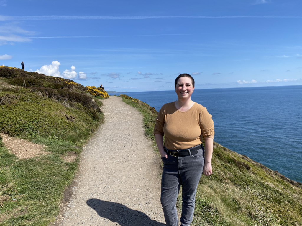 Ilana, a non-binary person wearing a tan long sleeved shirt and black jeans, smiles as they stand on a path near the ocean.